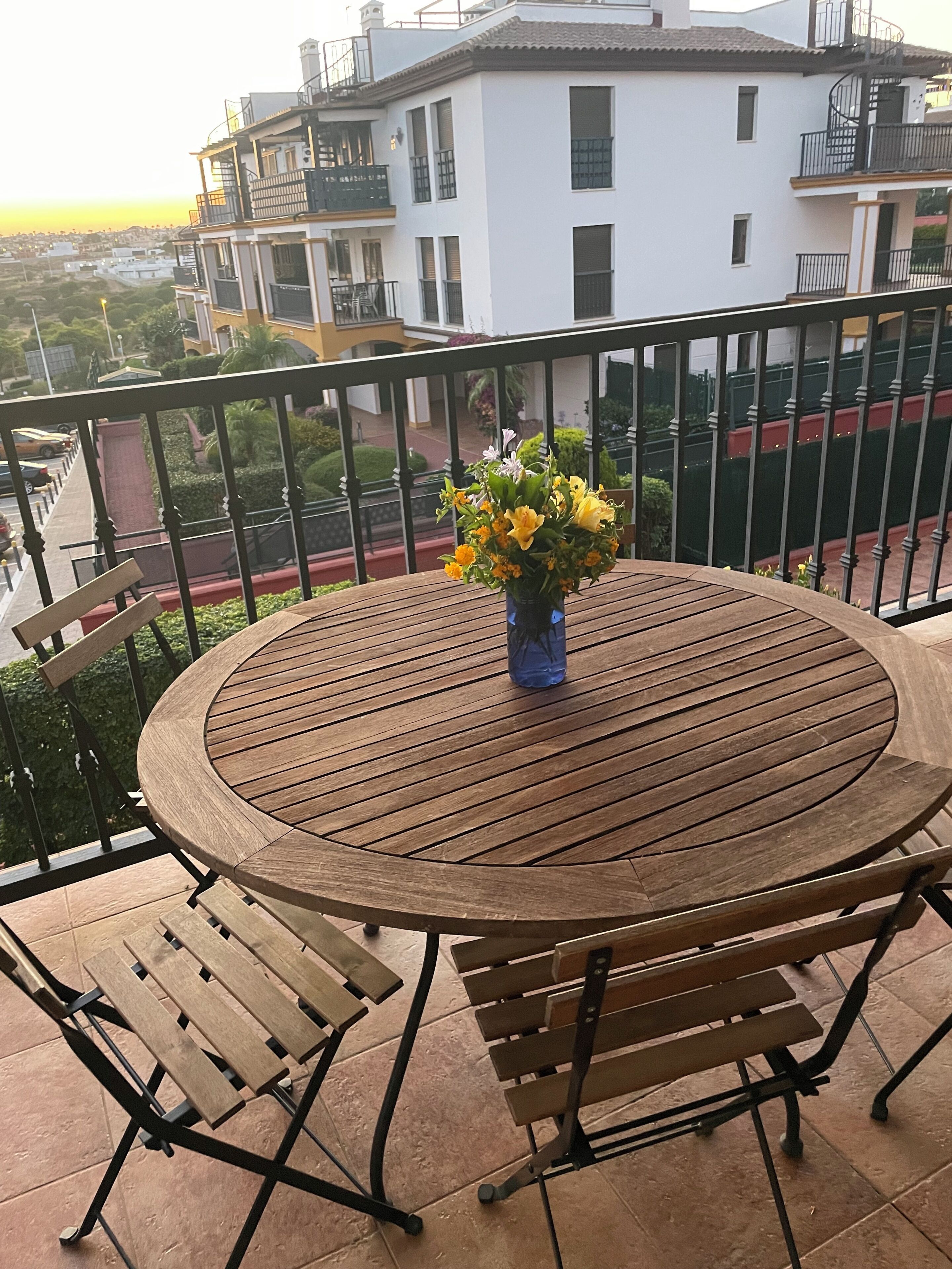 A round wooden table with a vase of flowers on a balcony with a metal railing.