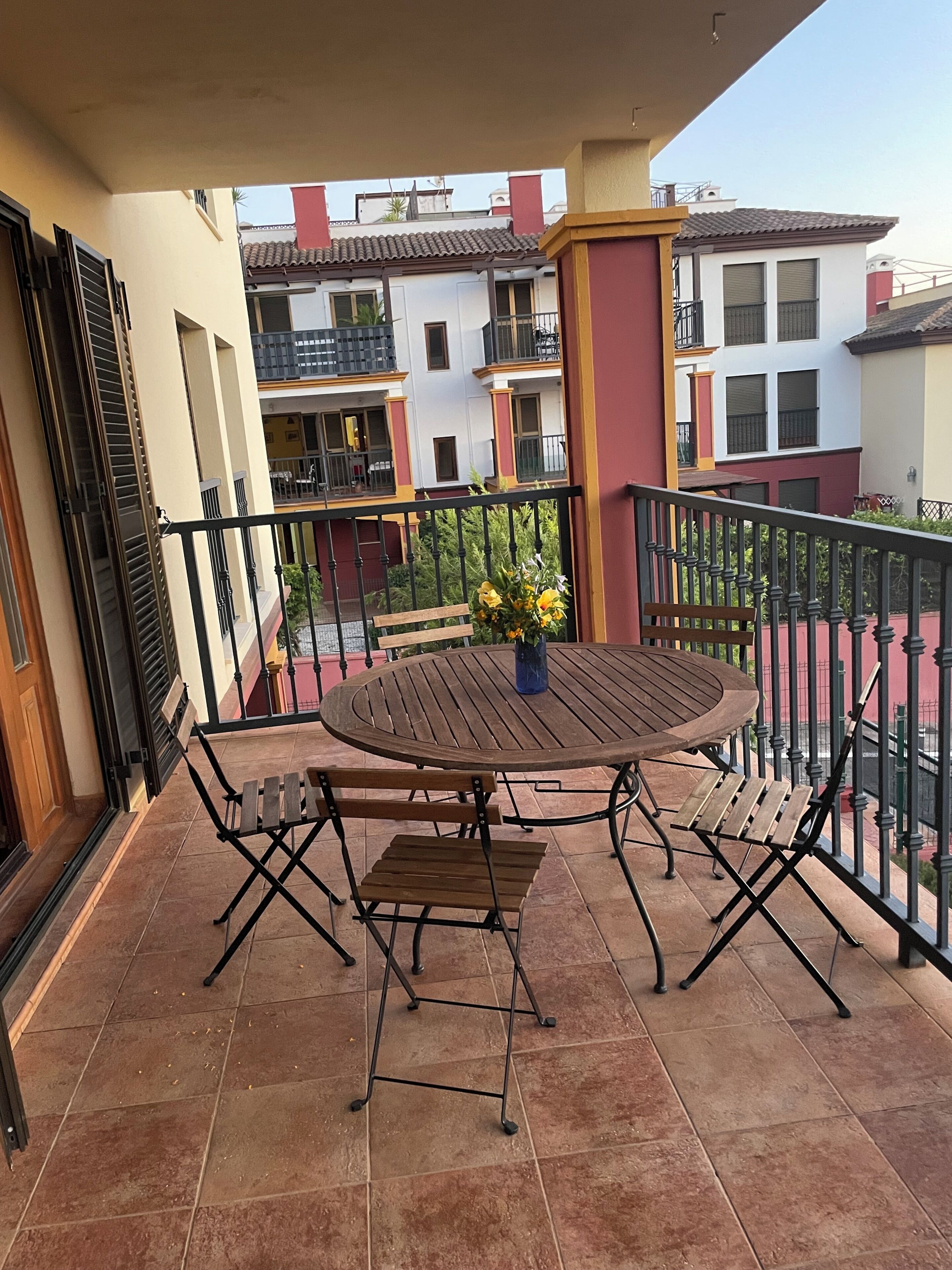 Un balcon avec une table ronde en bois et des chaises, un vase rempli de fleurs et une vue sur les bâtiments voisins.