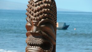 Een houten sculptuur met gedetailleerde houtsnijwerken, geplaatst aan het strand met een boot op de achtergrond.