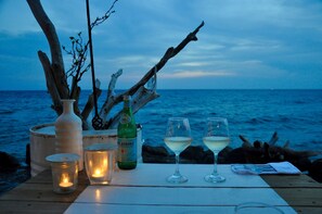 Una mesa con una botella de vino, dos copas, una vela y un florero junto al mar.