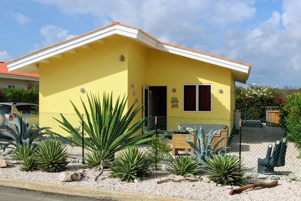Un edificio amarillo con techo rojo, un jardín cercado con plantas de agave y un banco de madera.