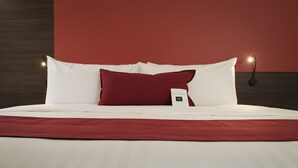 Ein ordentlich bezogenes Bett mit weißer und roter Bettwäsche, einem roten Kopfkissen und einem kleinen Nachttischchen mit einer Lampe.