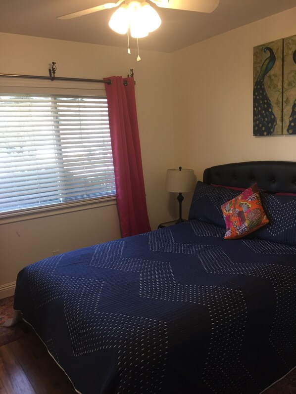 Une chambre à coucher avec un lit, une table de chevet, une lampe, une fenêtre avec des stores, un ventilateur de plafond et un tableau accroché au mur.