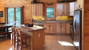Een houten keuken in een chalet, met een centraal eiland, houten kastjes, een roestvrijstalen koelkast en een raam met uitzicht op het bos.