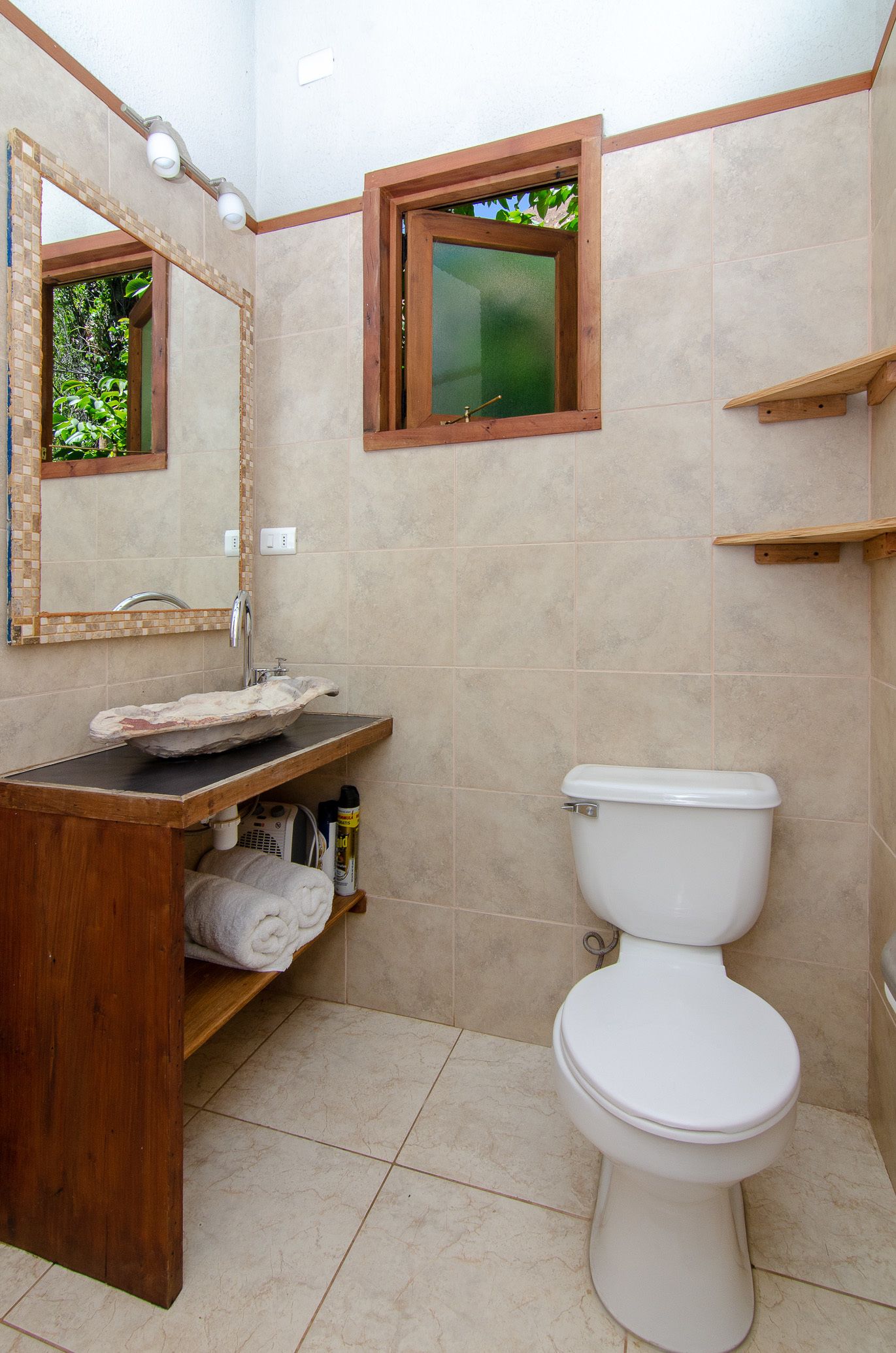 Un baño con tocador de madera, lavamanos blanco, inodoro y ventana con vistas a follaje.