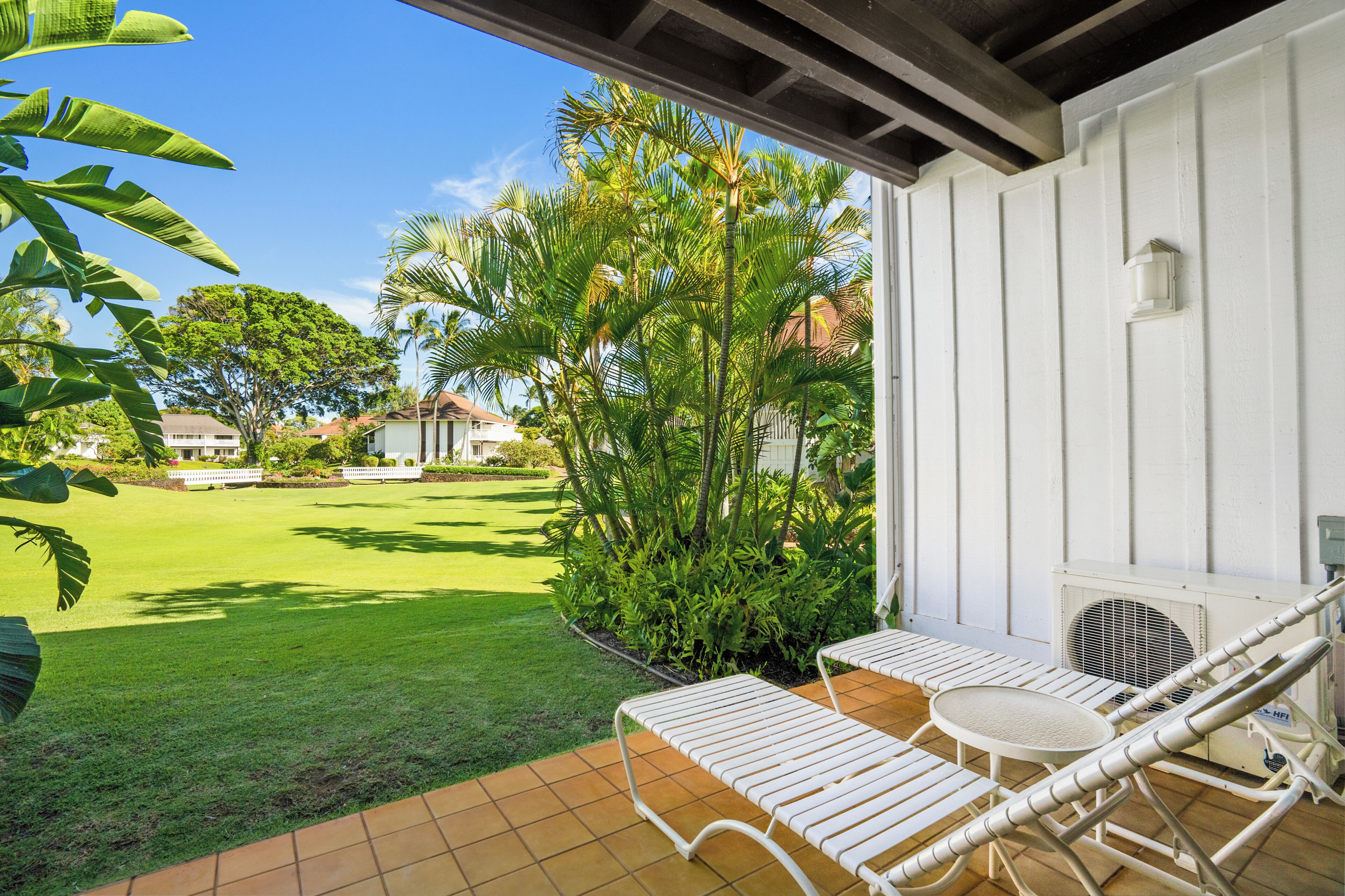 Un patio con dos sillones blancos y una mesita, rodeado de césped cuidado y plantas tropicales.