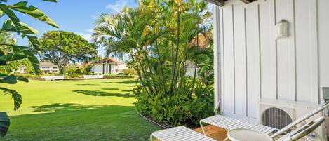 Un patio con dos sillones blancos y una mesita, rodeado de césped cuidado y plantas tropicales.