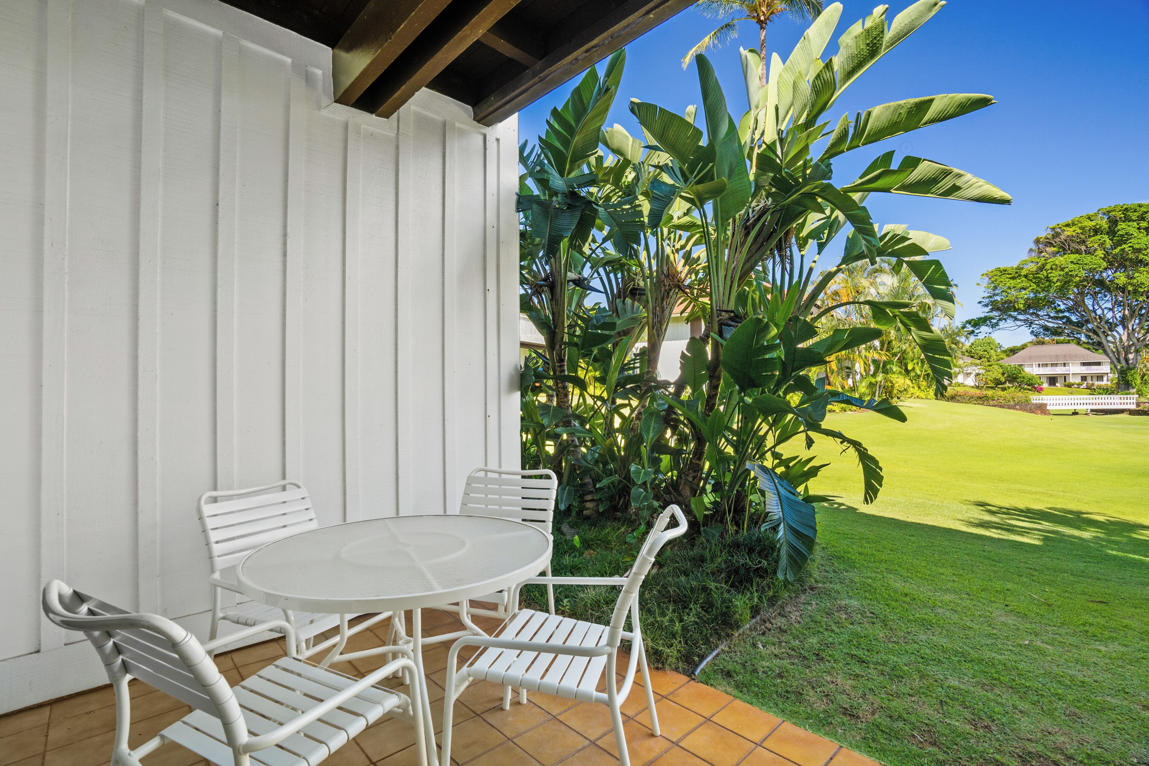 Une terrasse avec une table et des chaises blanches, entourée d’une végétation luxuriante et d’une pelouse bien entretenue.