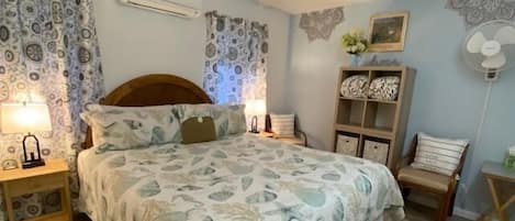 Un dormitorio con cama, mesitas de noche, una silla, una pequeña mesa y un ventilador de pared.