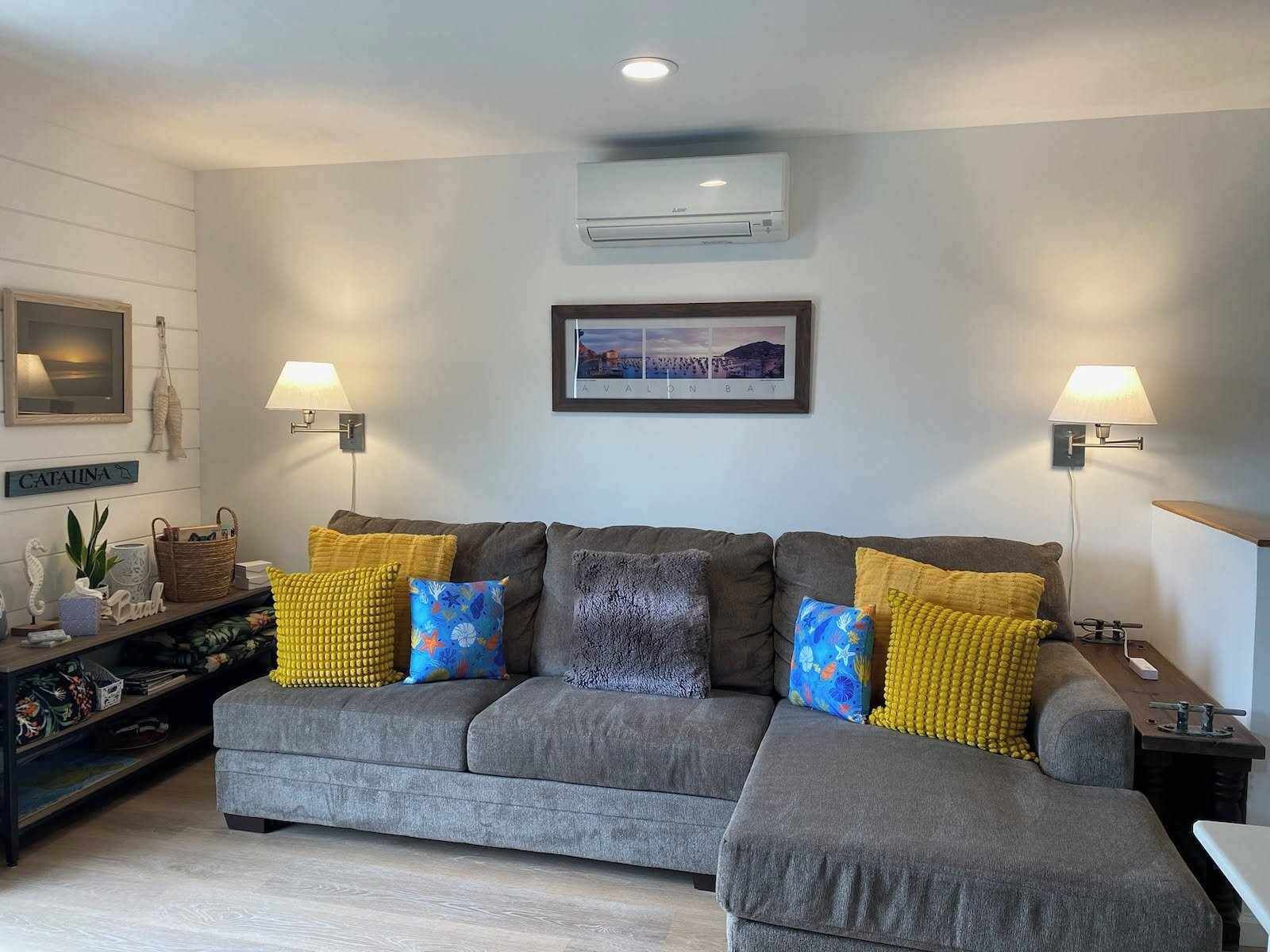 Una sala de estar con un sofá seccional gris, dos lámparas fijadas a la pared, un equipo de aire acondicionado, un cuadro enmarcado y un estante con adornos.