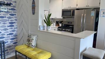 Cozinha moderna com armários brancos, geladeira de aço inoxidável, micro-ondas e fogão. Há um banco amarelo com almofada floral, uma planta em vaso e uma placa de boas-vindas na parede.