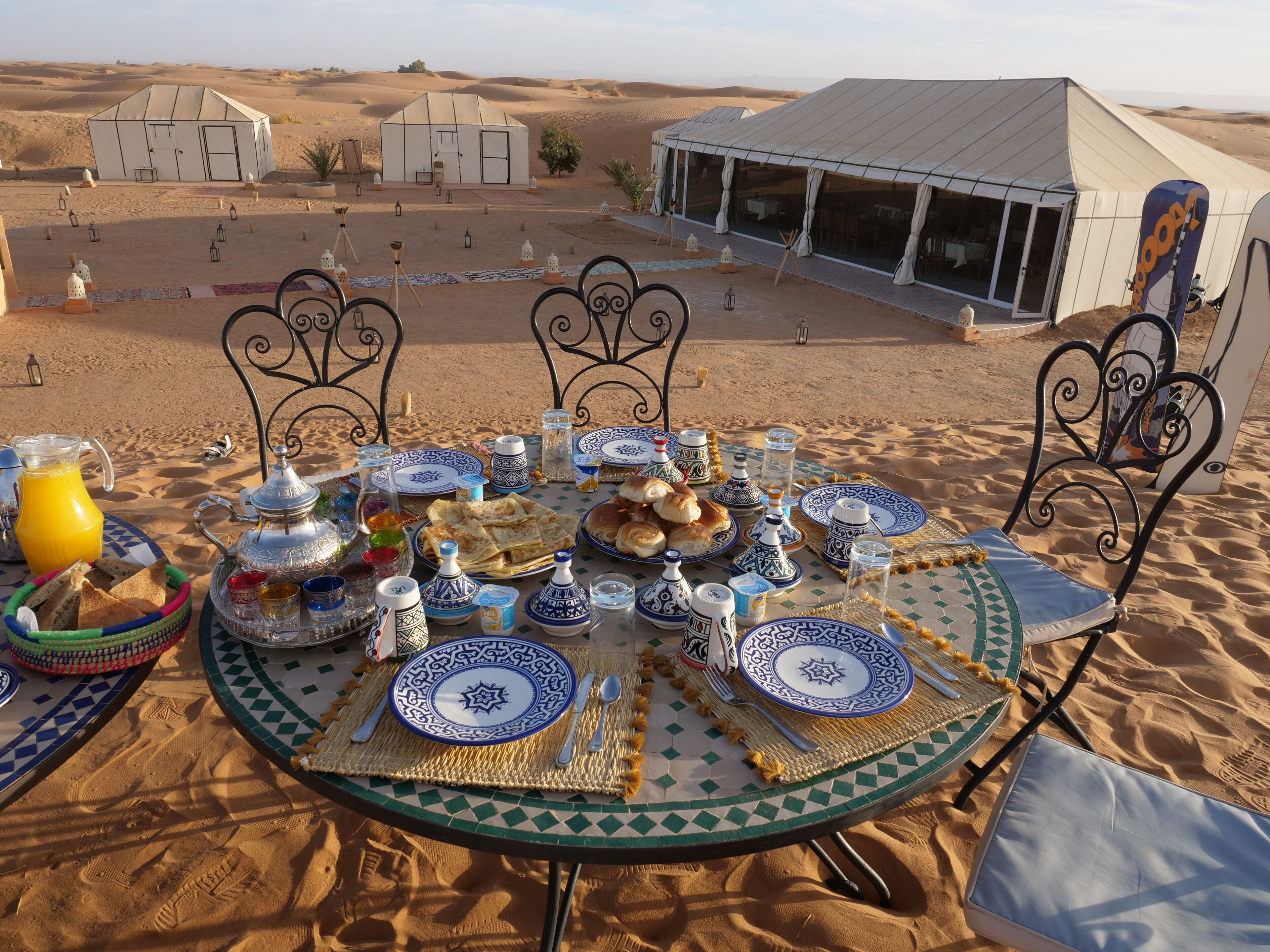 Un campamento en el desierto con una mesa dispuesta para una comida, rodeado de tiendas de campaña y un amplio espacio abierto.