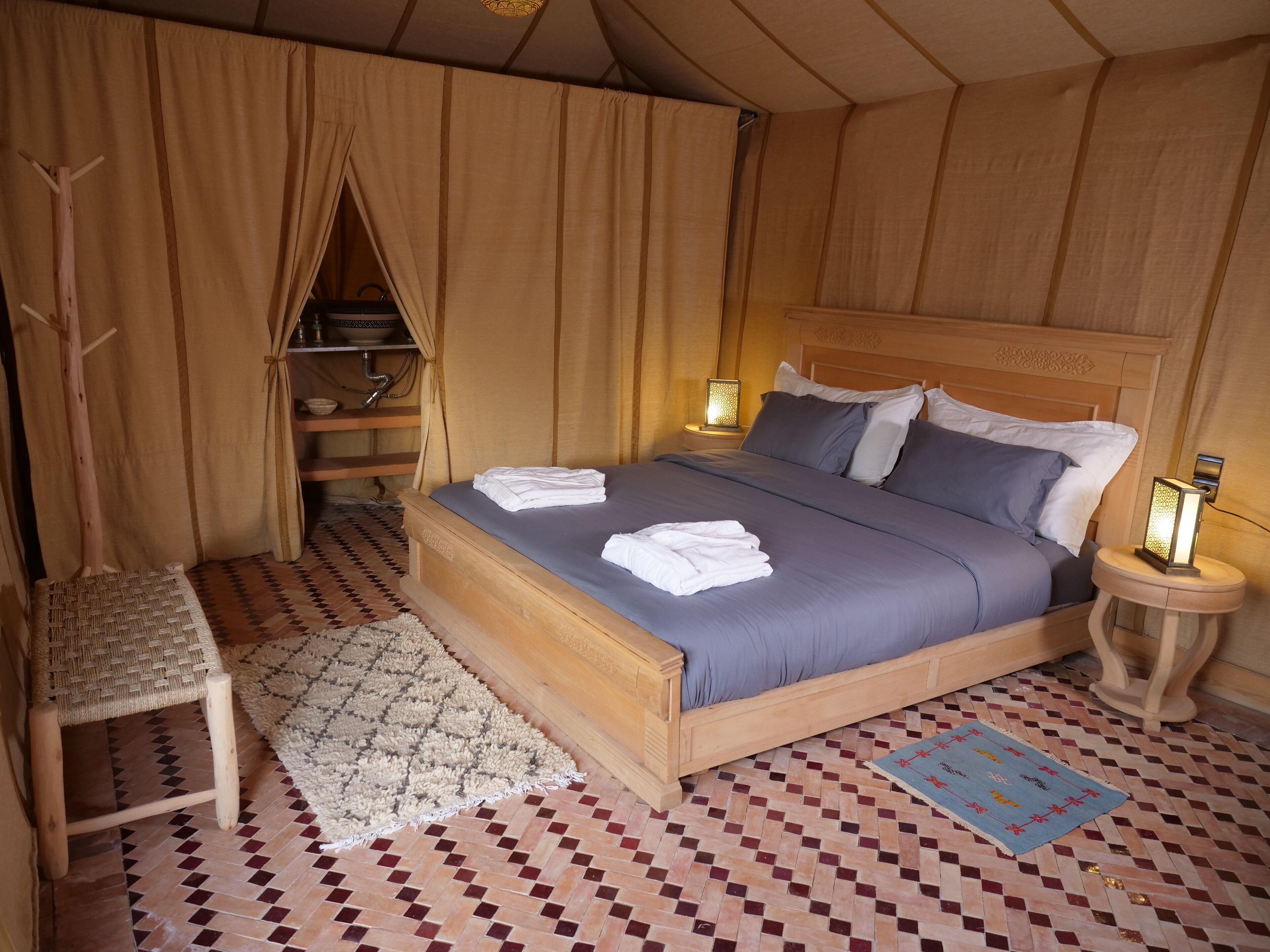 A neatly made bed with a headboard, a bedside table with a lamp, a chair, and a patterned rug on a tiled floor.