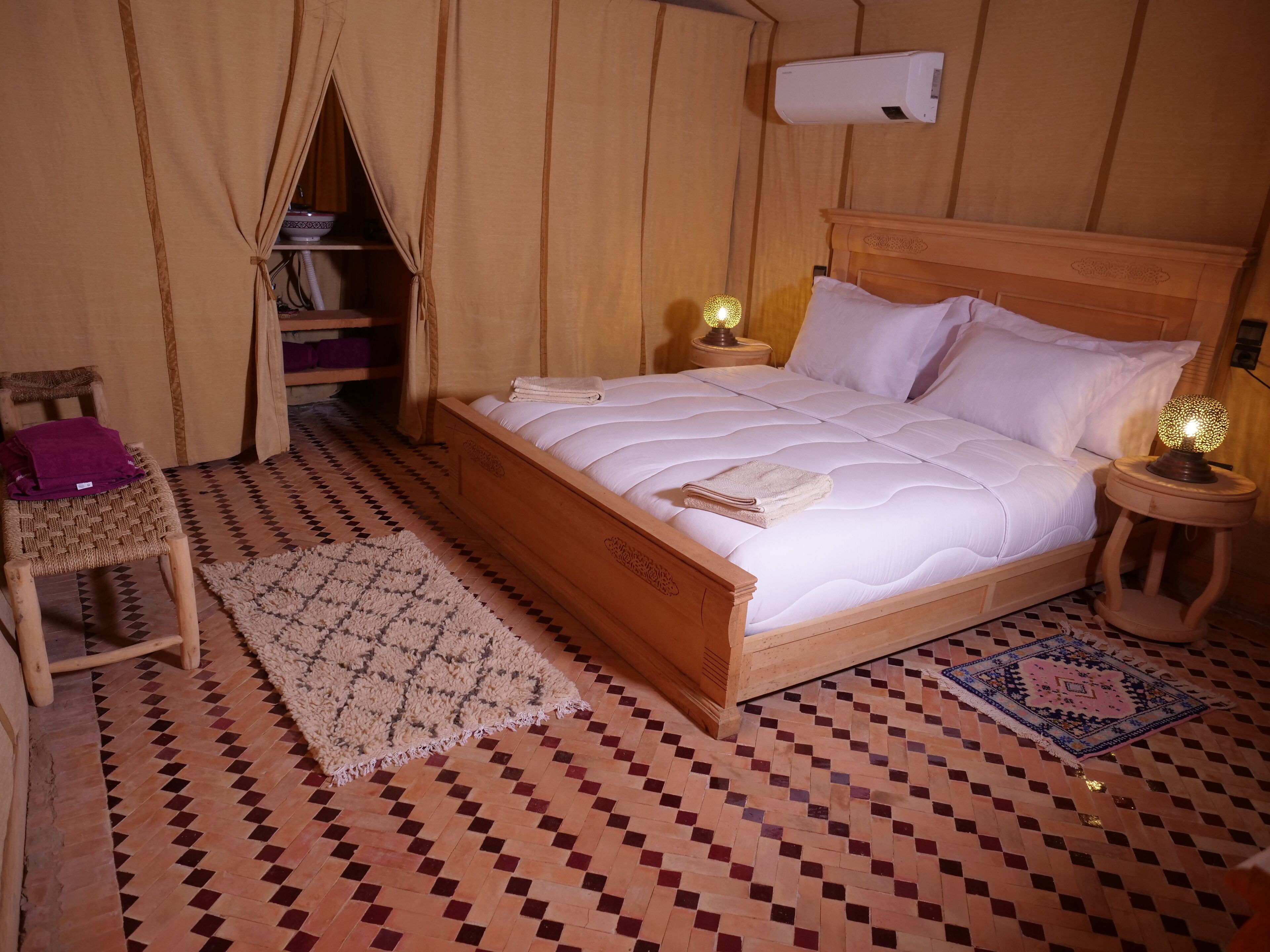 A wooden bed with white bedding, two bedside lamps, a small wooden chair, a patterned rug, and a shelf with folded towels.