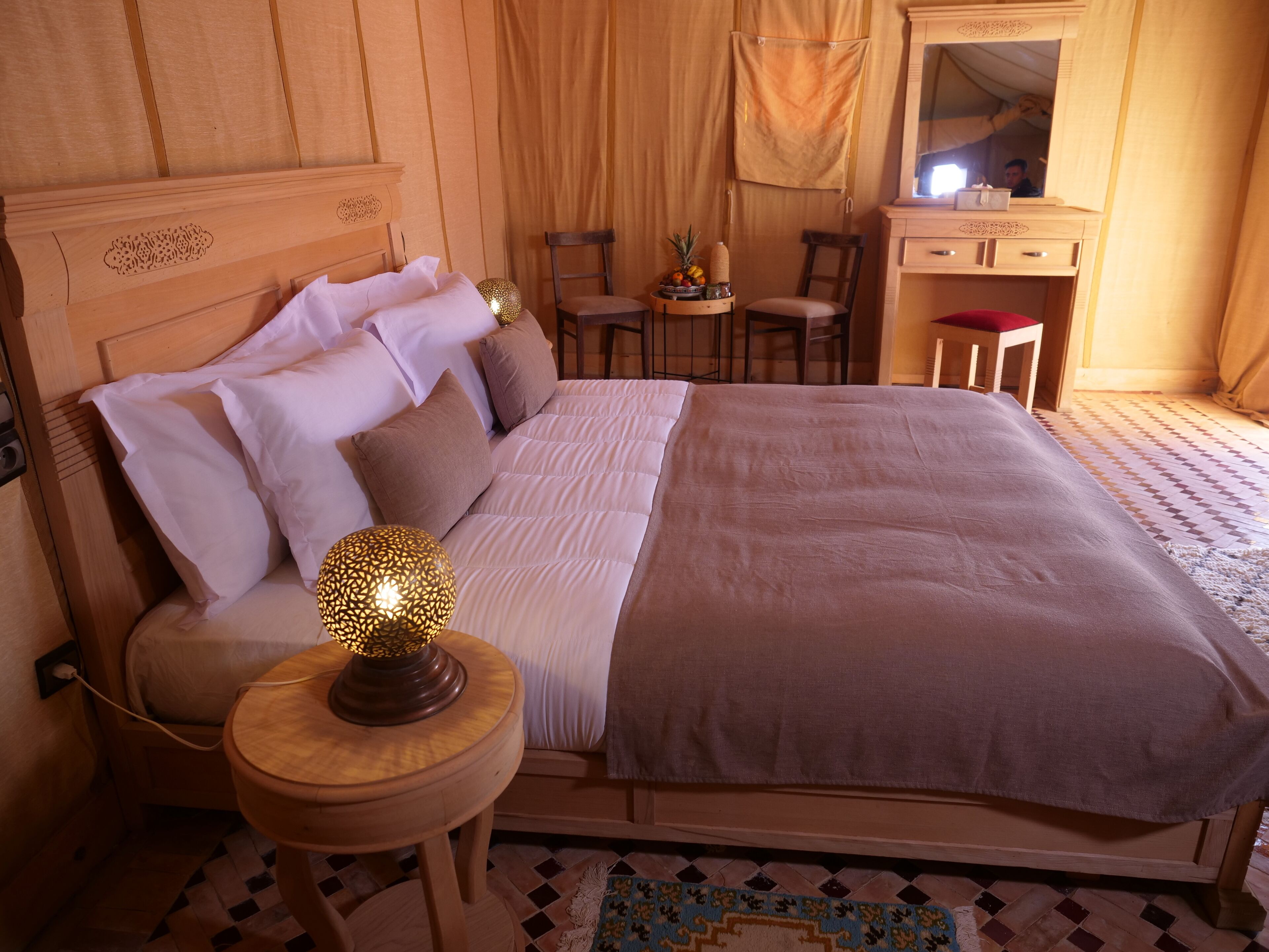 Un dormitorio ordenado con cama, lámparas de noche, una mesita con fruteros, una silla y un espejo.