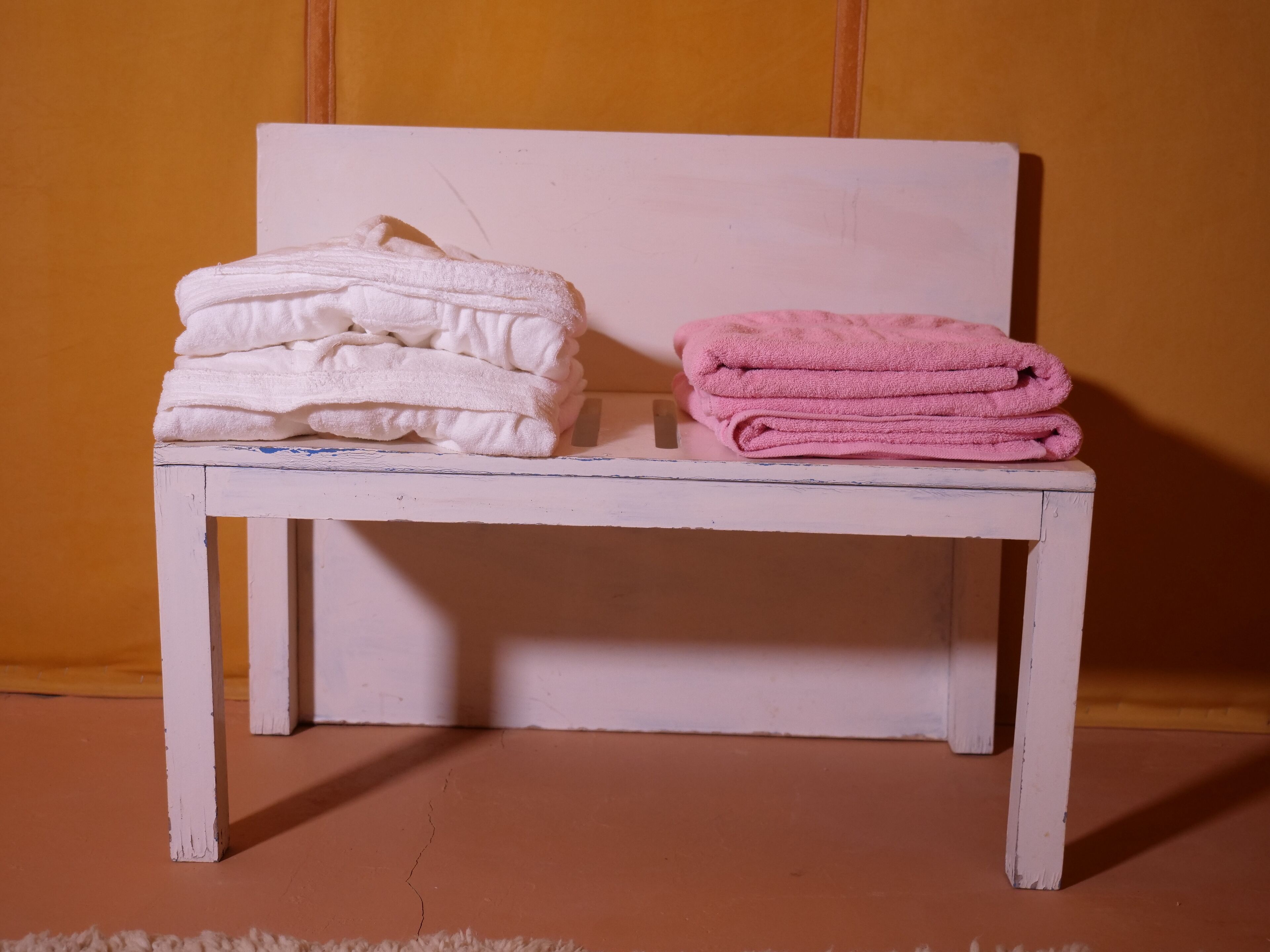A small white bench with two stacks of folded towels, one white and one pink, against a plain wall.