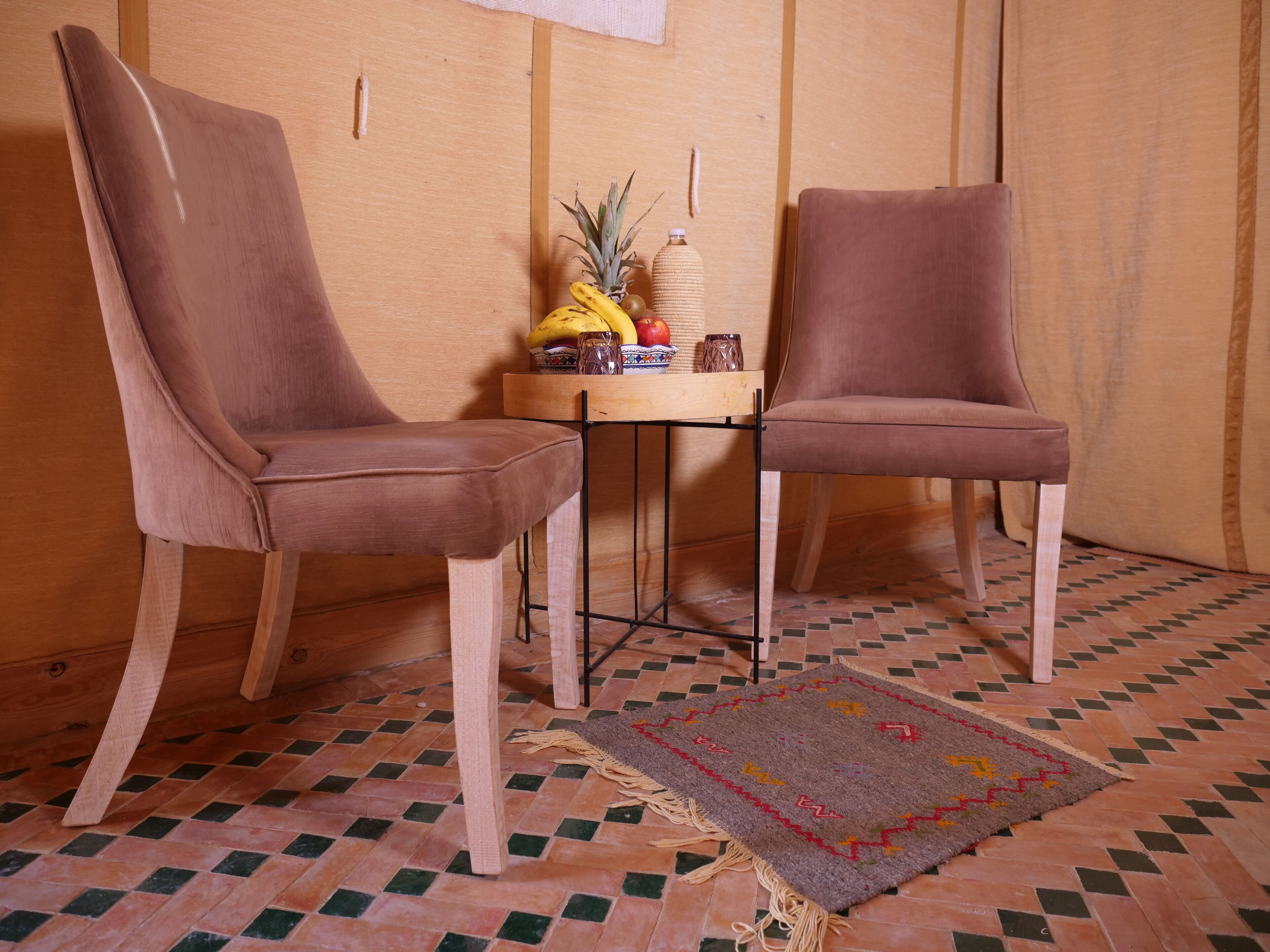 Two brown upholstered chairs with wooden legs, a small round table with a woven basket, a pineapple, and a bowl of fruit.