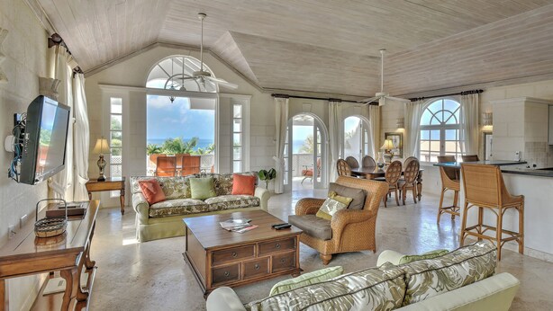 A spacious living area with a high ceiling, a dining area with wicker furniture, and a kitchen with a bar.