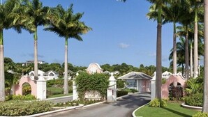 A gated entrance with palm trees, a curved road, and a building with a pink facade in the background.