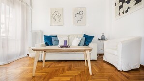 A living room with a wooden herringbone floor, a white sofa with blue cushions, a small side table with a lamp, and two framed artworks on the wall. - Acropolis unique view (Athina)