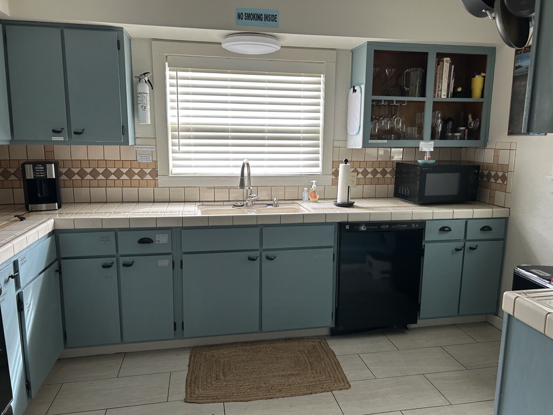 A kitchen with light blue cabinets, a sink, a microwave, and a window with blinds.