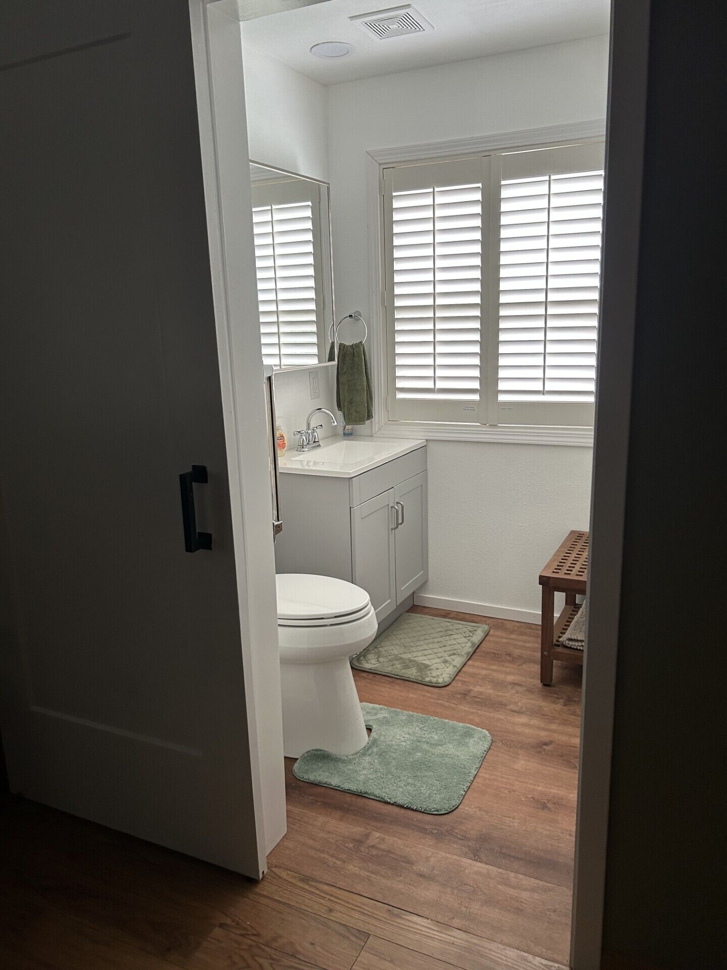 A bathroom with a toilet, sink, and window with blinds. There is a wooden bench and a green rug on the floor.