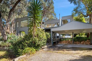 Una casa moderna de dos pisos con un patio cubierto y un jardín con diversas plantas y árboles.