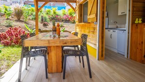 Um deck de madeira com uma mesa e cadeiras, uma área de cozinha com armários e pia, e um jardim com diversas plantas e flores.