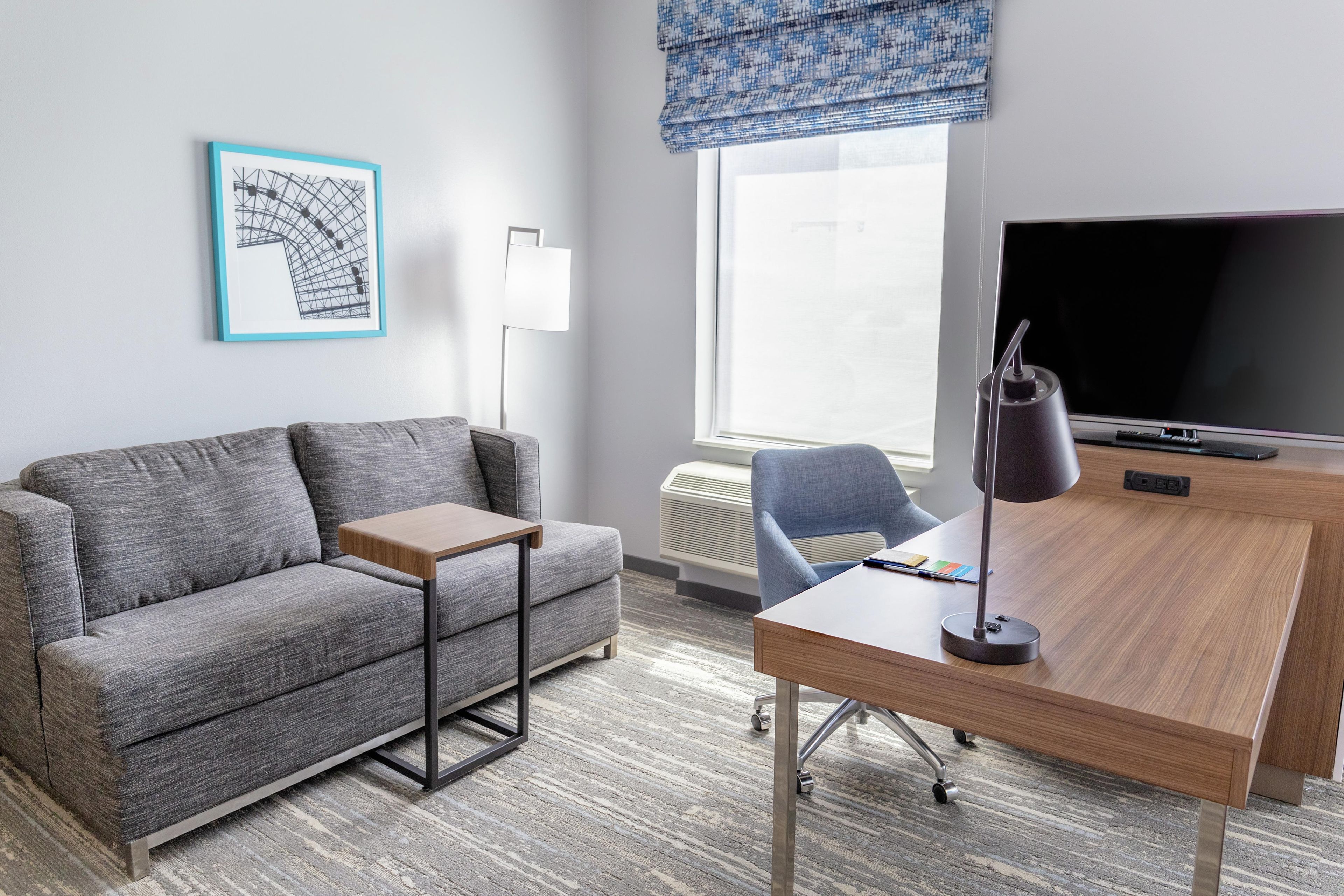 Una habitación de hotel moderna con un sofá gris, una pequeña mesita de madera, un escritorio con una silla, un televisor de pantalla plana grande y una ventana con un visillo azul.