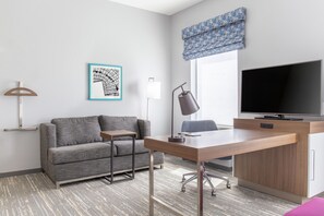 A modern hotel room with a grey sofa, a wooden desk, a flat-screen TV, and a patterned window valance. - Hampton Inn & Suites Bridgeview Chicago (Summit)