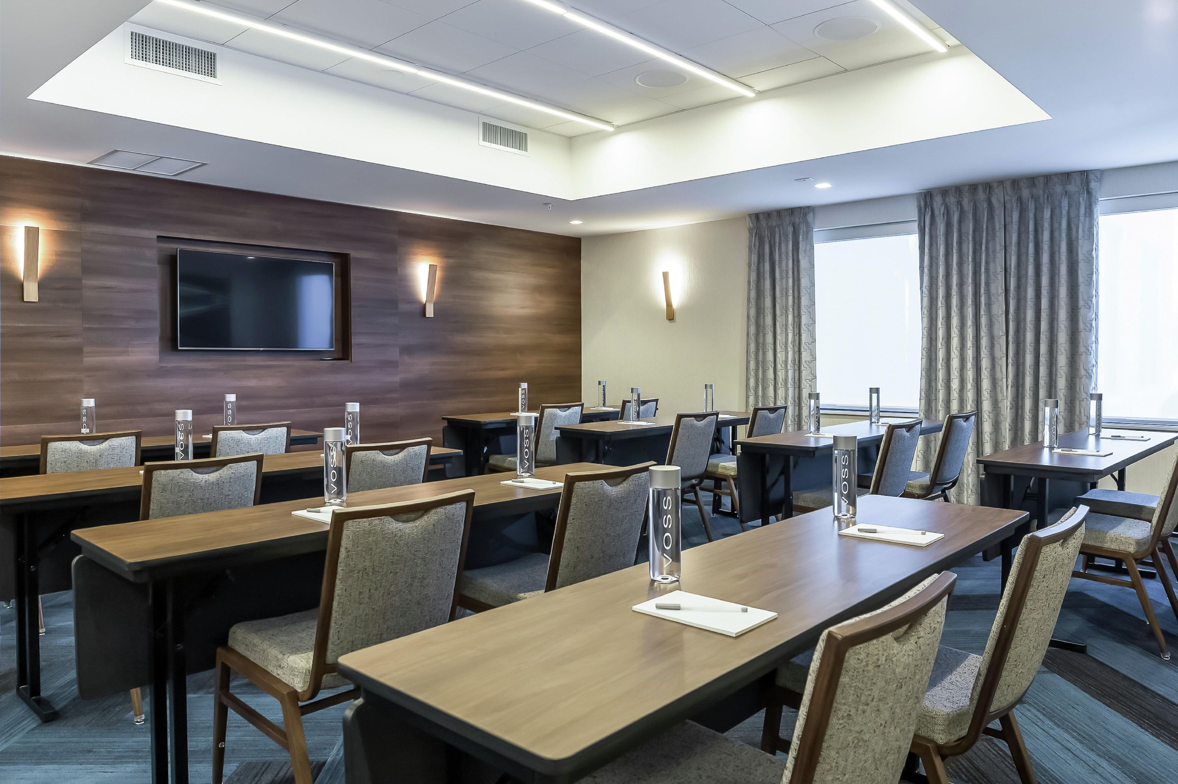 A conference room with multiple tables and chairs, equipped with water bottles and a flat-screen TV mounted on the wall.
