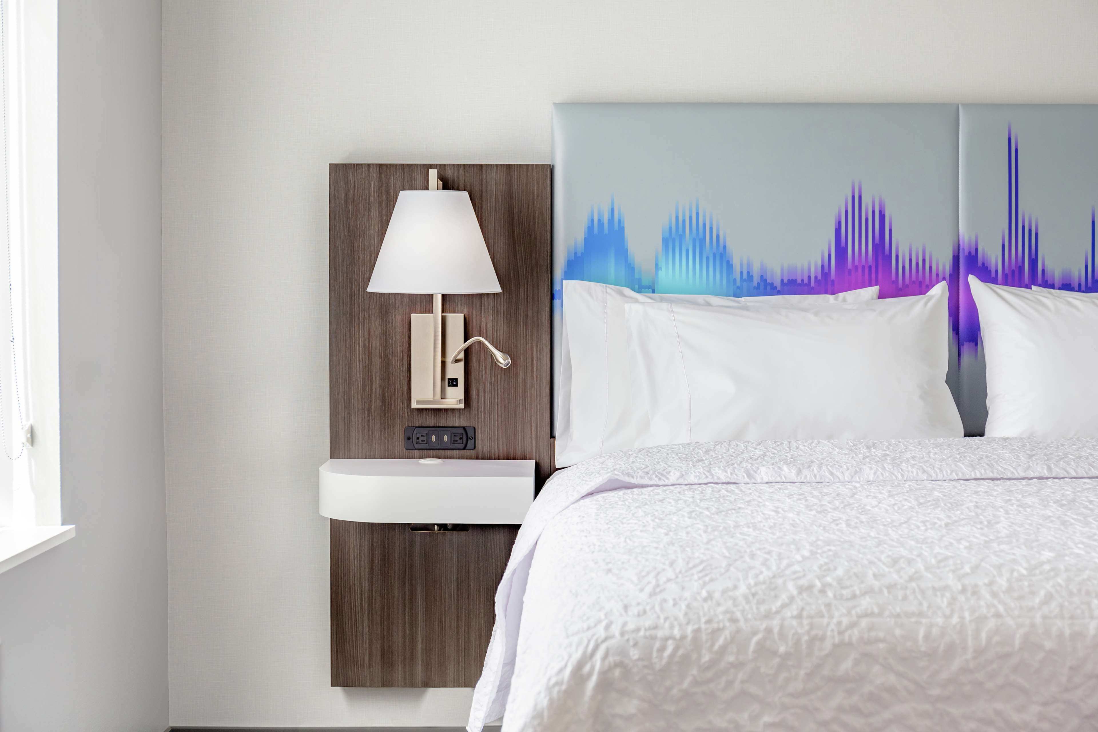 A hotel room with a bed, a bedside table with a lamp, and a wall-mounted sink.