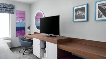 Ett modernt hotellrum med ett skrivbord i trä, en platt-TV, en grå kontorsstol, en lila bänk och konstverk på väggarna.