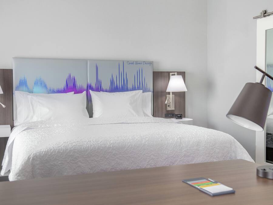 A hotel room with a bed, two bedside tables, a nightstand, a lamp, and a smartphone on a wooden table.