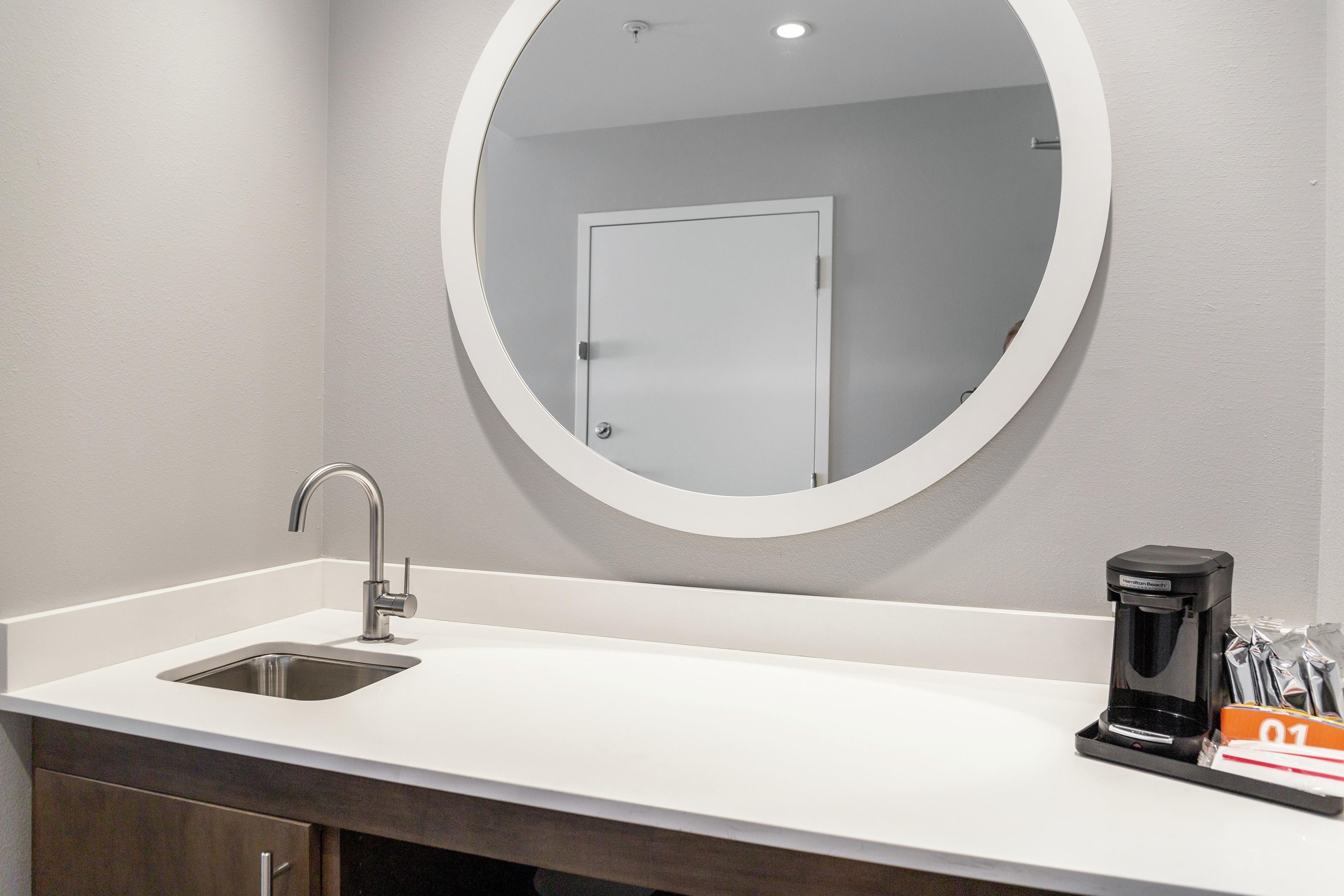 Un baño moderno con un lavamanos blanco, un espejo redondo y una cafetera sobre el mostrador.