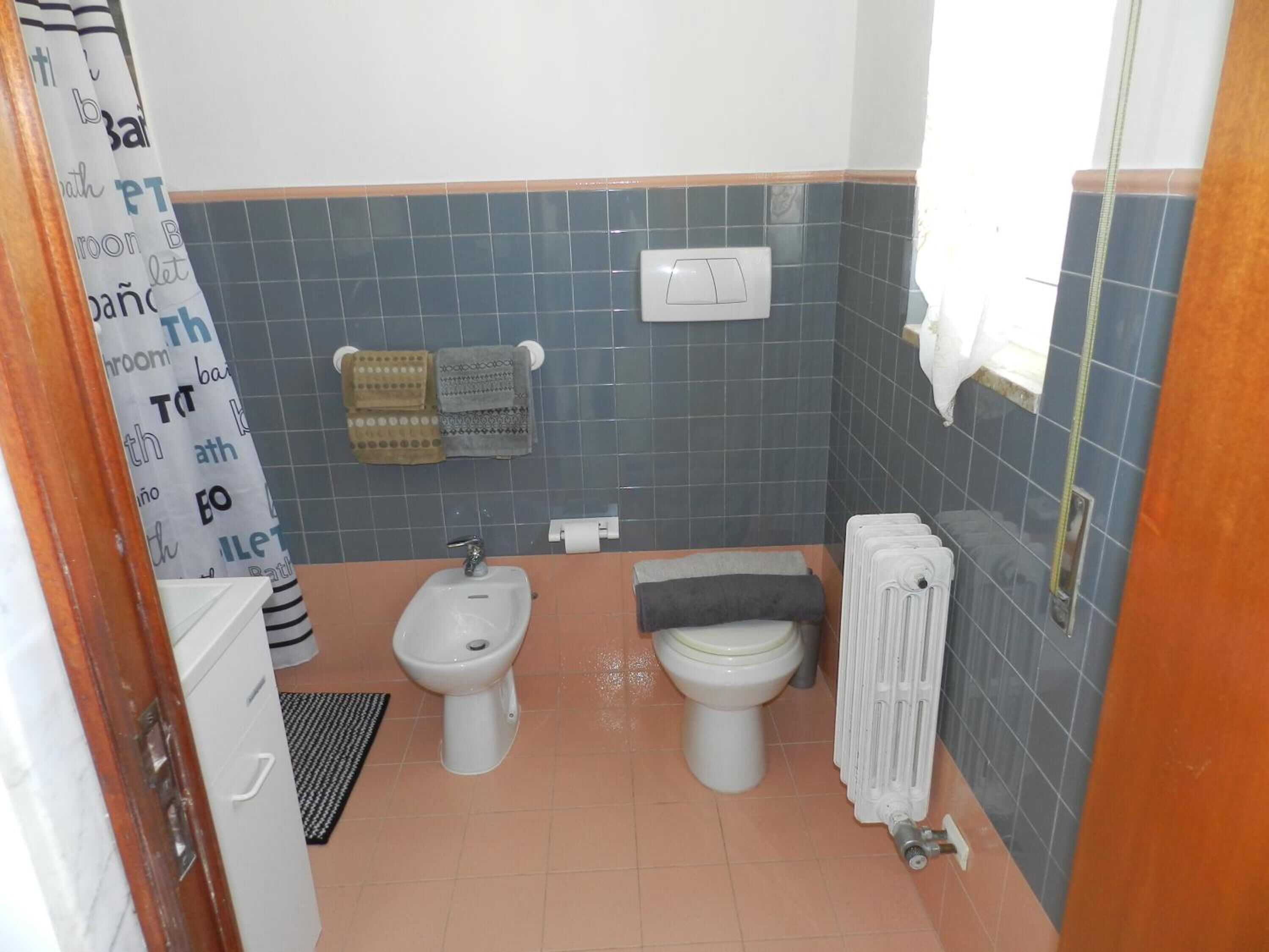 A bathroom with two toilets, a bidet, a towel rack with towels, a radiator, and a shower curtain with text.