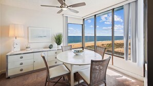Habitació amb taula de dinar, cadires, ventilador de sostre, finestra gran amb vista a la platja i un armari blanc amb calaixos.