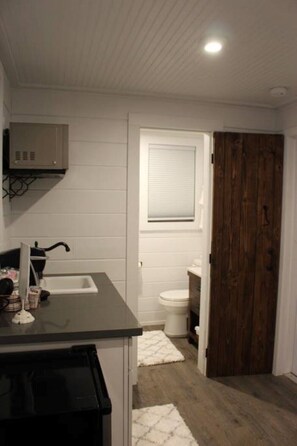 Ein Badezimmer mit einem weißen Waschbecken, einer Toilette und einem Fenster. Eine Holztür und ein weißer Wandheizer sind ebenfalls vorhanden.