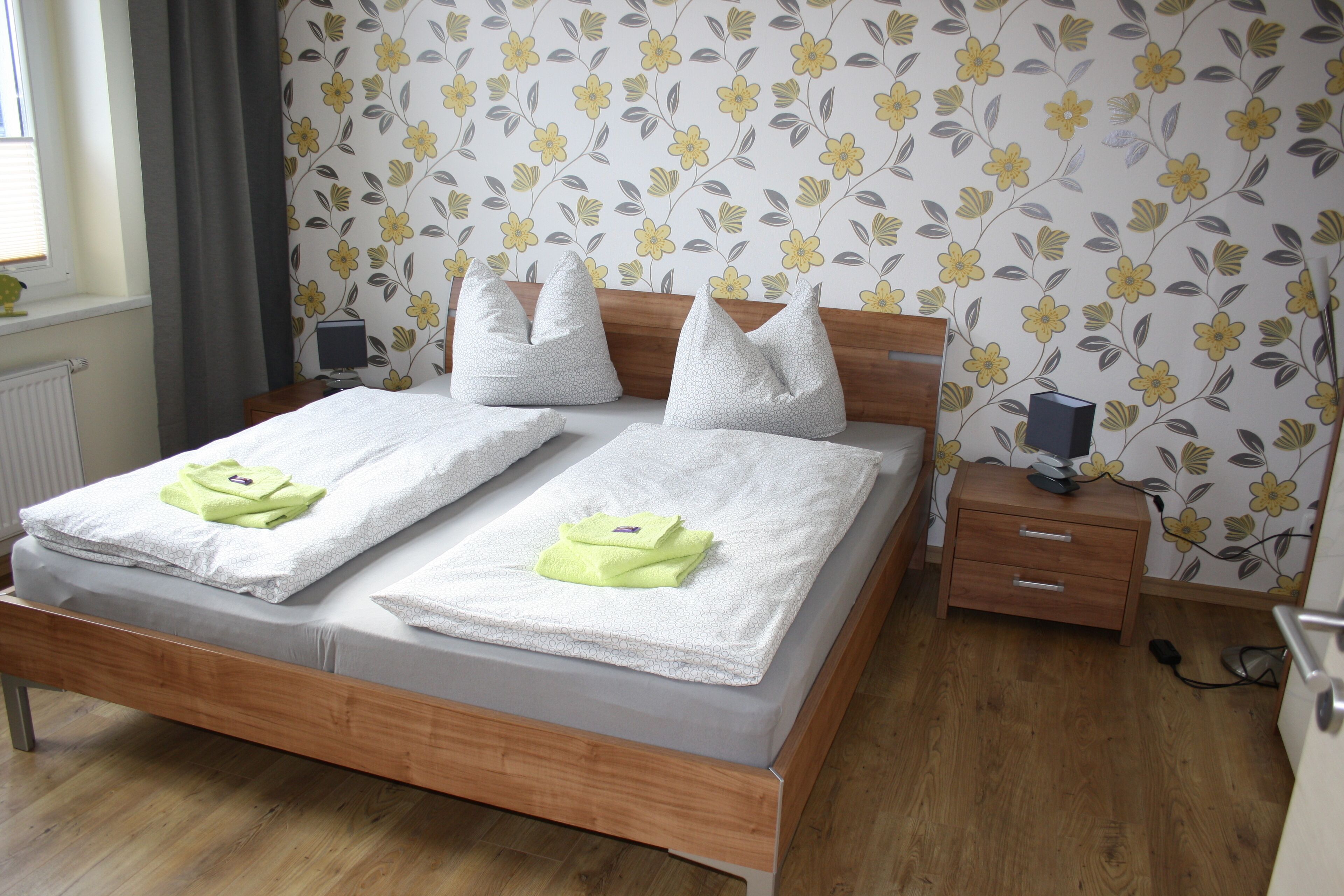 A neatly made bed with two pillows and two folded towels, a wooden bedside table with a lamp, and a floral wallpaper pattern on the wall.