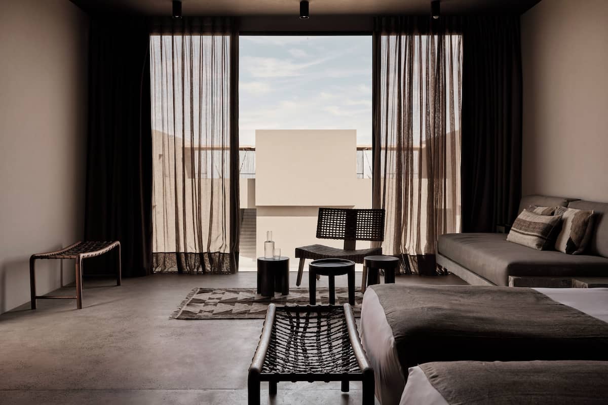 Habitación de hotel moderna con una cama grande, un sofá, una mesita pequeña y una silla. Se ve el cielo a través de las cortinas.