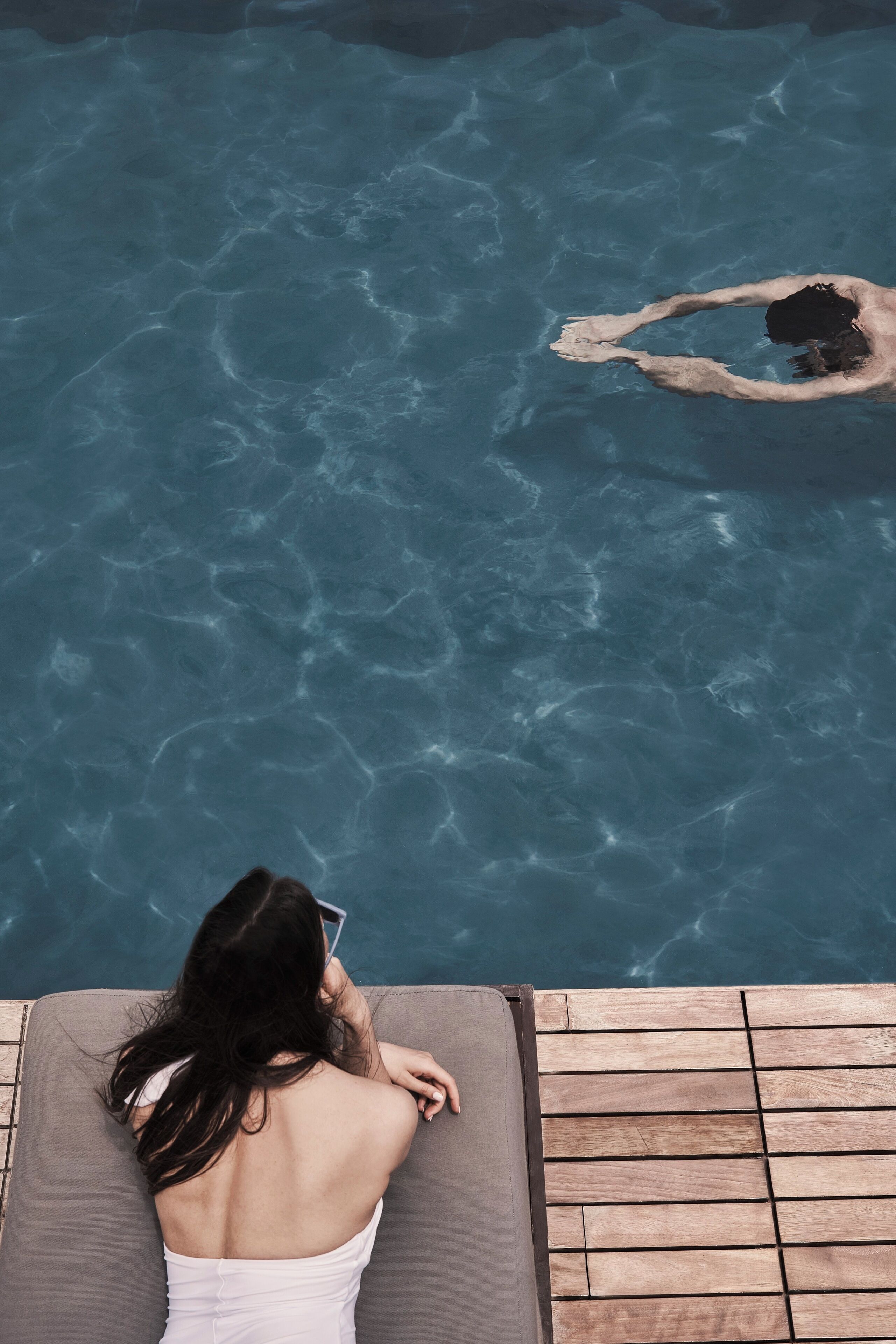 A person relaxing on a wooden deck by a clear pool with a small island.