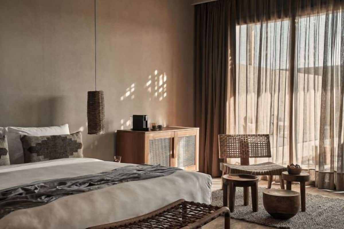 Un dormitorio con cama, un armario de madera, una silla de mimbre, una mesita pequeña y un amplio ventanal con cortinas.