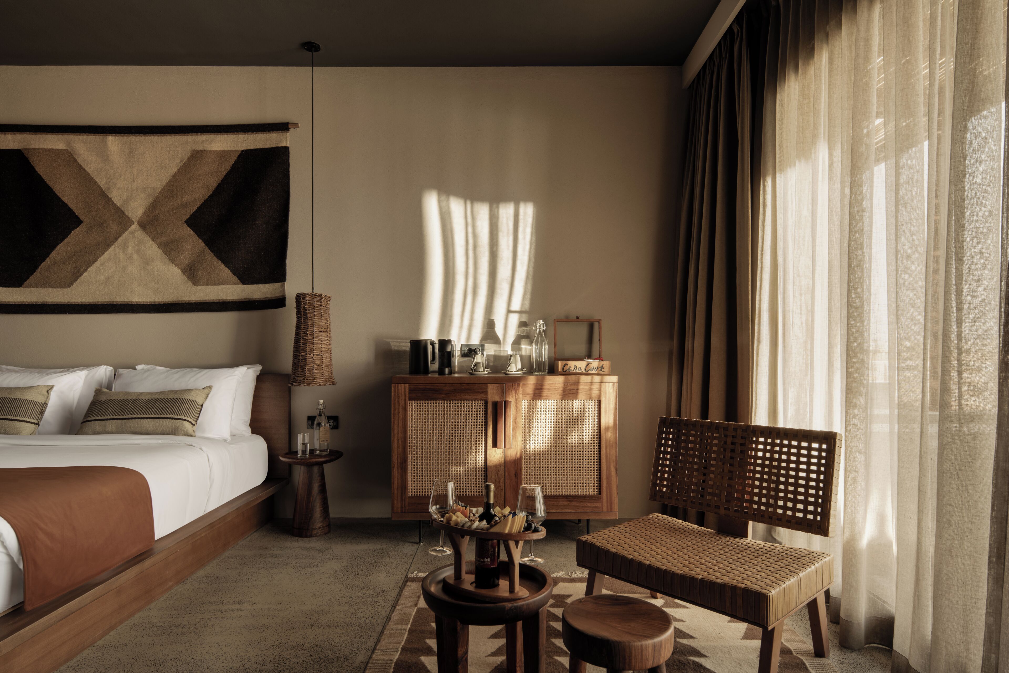 Een hotelkamer met een bed, een houten bankje, een klein tafeltje met een vaas, een rieten bijzettafeltje en een kast met een spiegel.