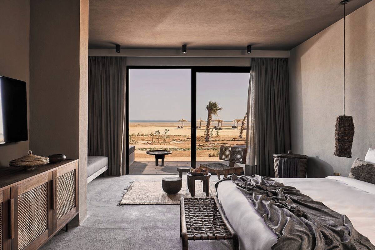 Habitación de hotel con una cama grande, televisión, consola de madera, balcón con vistas a la playa y una silla de mimbre.