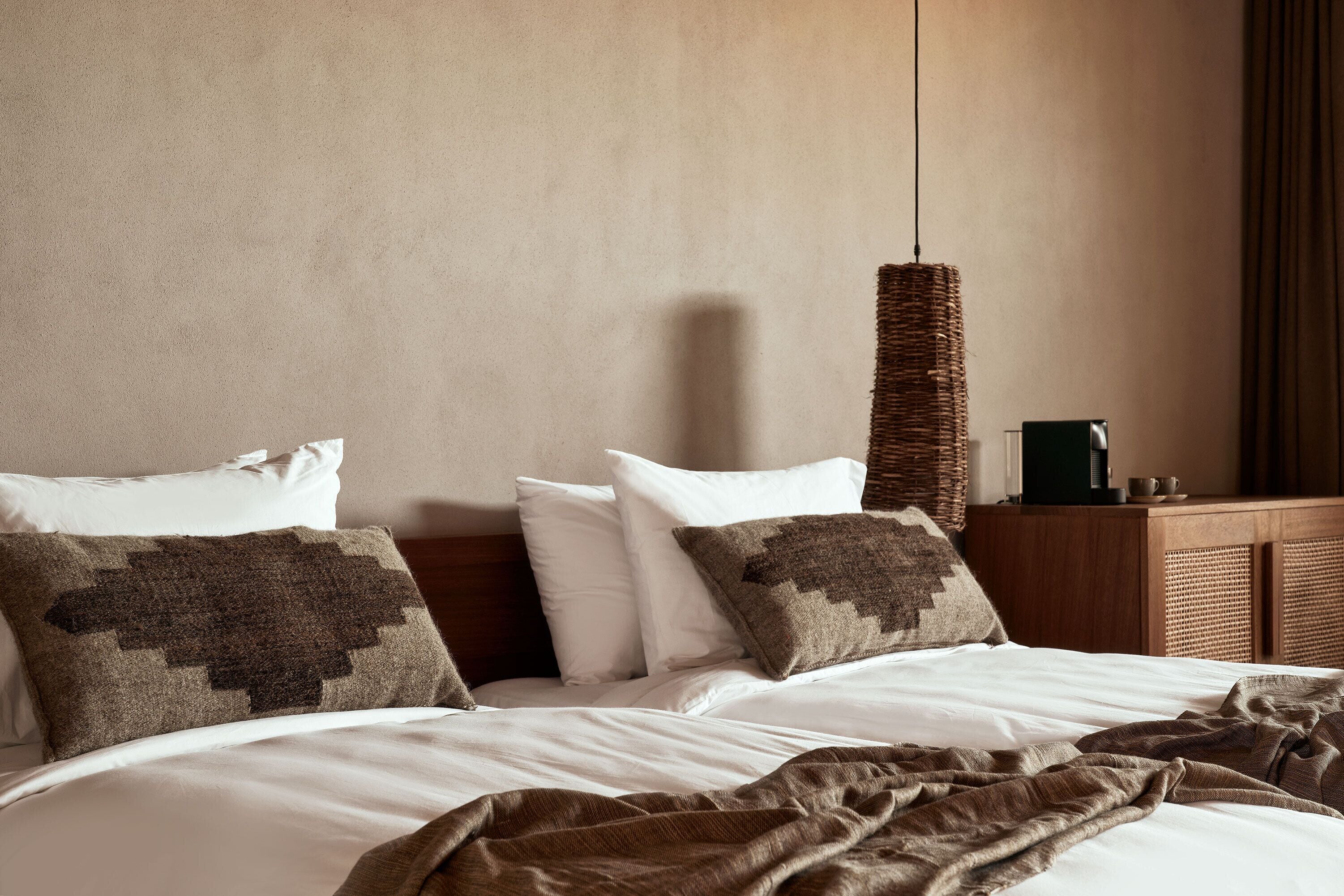 A neatly made bed with white linens and a brown blanket, a woven wall hanging, and a wooden nightstand with a cup and a small object on it.