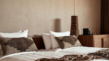 A neatly made bed with white linens and a brown blanket, a woven wall hanging, and a wooden nightstand with a cup and a small object on it.