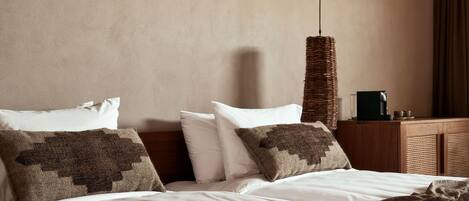 A neatly made bed with white linens and a brown blanket, a woven wall hanging, and a wooden nightstand with a cup and a small object on it.