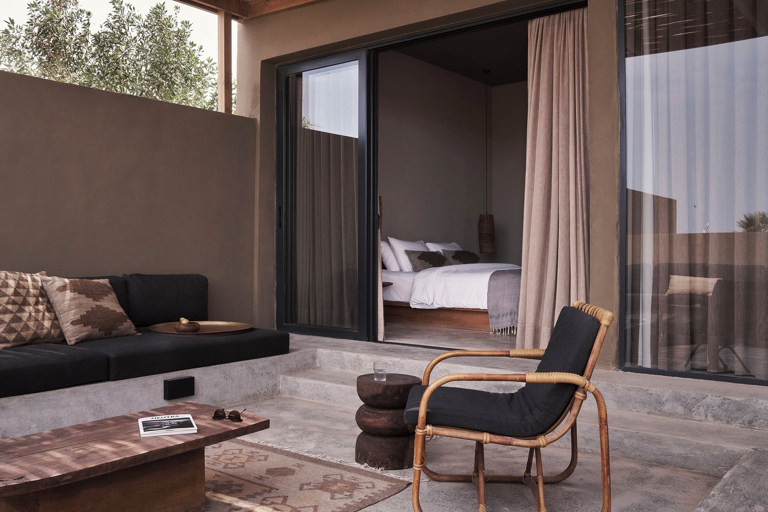 Un espace de vie moderne comprenant un canapé, une chaise en bois, une table basse et une vue sur une chambre à coucher, visible à travers des portes vitrées.