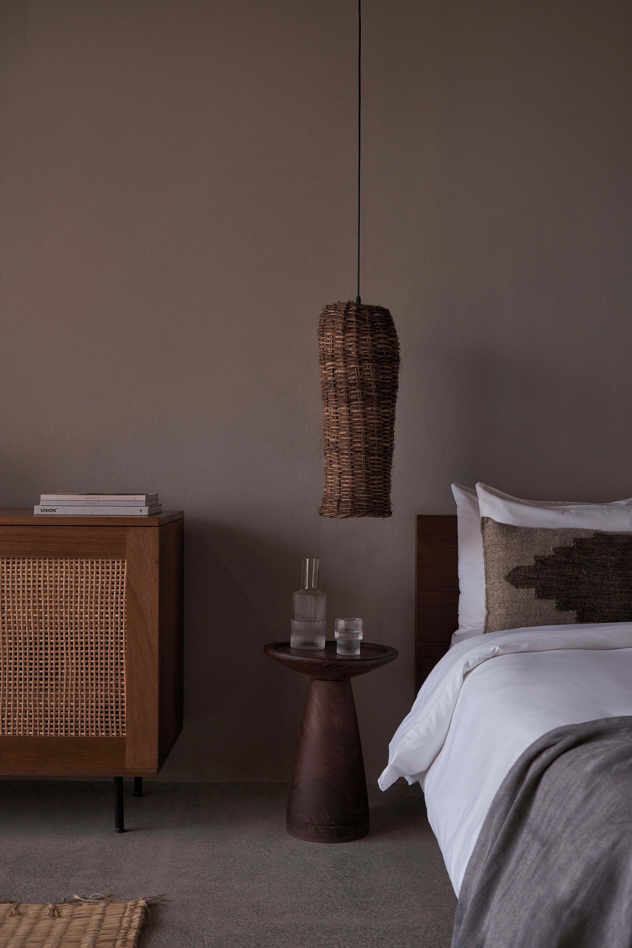Une chambre à coucher comprenant un lit, une table de chevet avec une lampe, une commode en bois et un luminaire suspendu en fibres tressées.