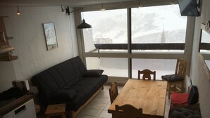 A cozy living area with a sofa, a wooden dining table, and chairs. There is a small kitchen area with a dishwasher and a refrigerator.